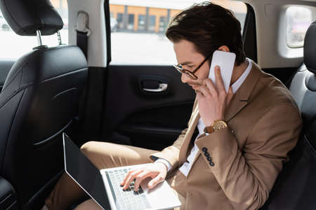 businessman in glasses working remotely with laptop while talking on smartphone in carの写真素材