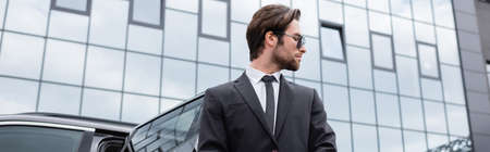 side view of bearded bodyguard in suit near car and modern building, bannerの写真素材
