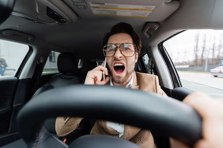 angry man talking on smartphone while driving car on blurred foregroundの写真素材