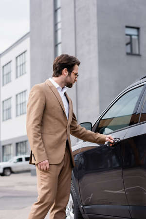 bearded man in beige jacket and glasses reaching car door