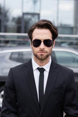 bearded bodyguard in suit and sunglasses with security earpiece near modern car on blurred...