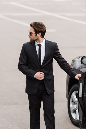 bearded bodyguard in suit and sunglasses with security earpiece opening door of modern auto