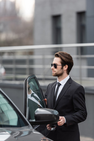 bearded man in suit and sunglasses standing near modern car with opened door