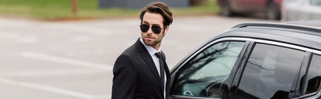 bearded bodyguard in suit and sunglasses with security earpiece near modern auto, banner