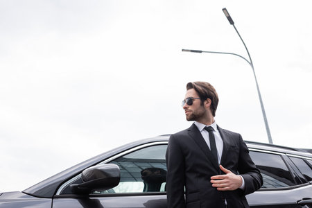 low angle view of bearded bodyguard in sunglasses and suit standing near modern carの写真素材