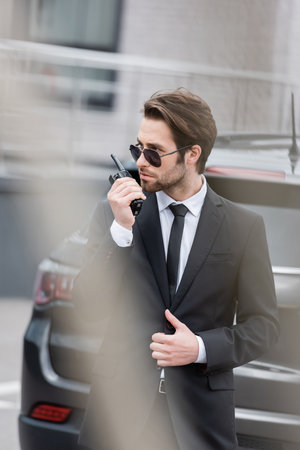 bodyguard in sunglasses and suit using walkie talkie near blurred modern car