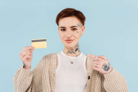 cheerful and tattooed woman pointing at finger at credit card isolated on blue