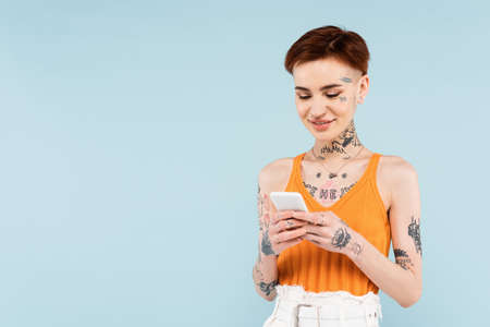 pleased and tattooed woman chatting on smartphone isolated on blue