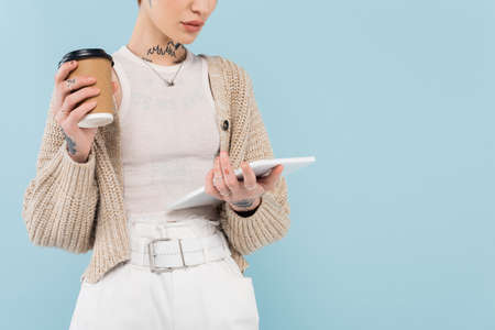 cropped view of young woman with tattoos holding digital tablet and paper cup isolated on blueの写真素材