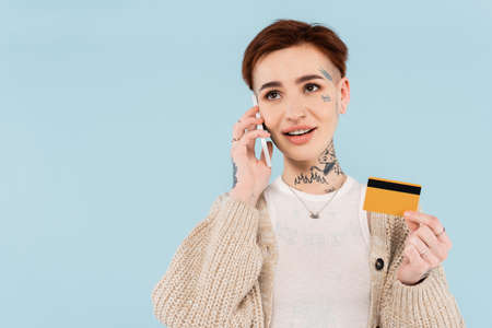 cheerful and tattooed woman holding credit card while making order on smartphone isolated on blueの写真素材