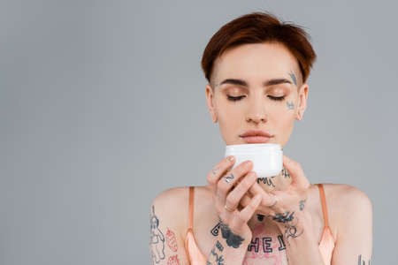 young woman with tattoos and red hair holding white container while smelling cosmetic product isolated on grayの写真素材