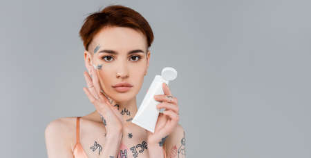 young woman with tattoos holding white tube and applying cream on face isolated on gray, bannerの写真素材