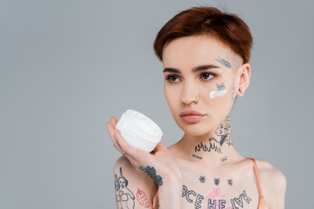 young woman with tattoos and red hair holding white container with cosmetic product isolated on grayの写真素材