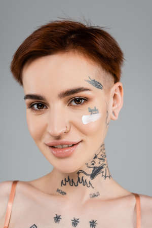 tattooed and happy woman with cosmetic cream on face smiling isolated on grayの写真素材