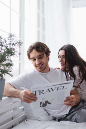Smiling man reading travel newspaper near girlfriend on bedの写真素材