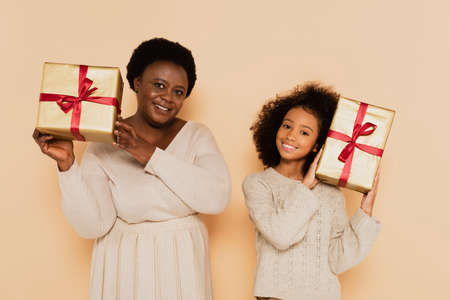 happy african american granddaughter and grandmother holding gift boxes near heads on beige backgroundの写真素材