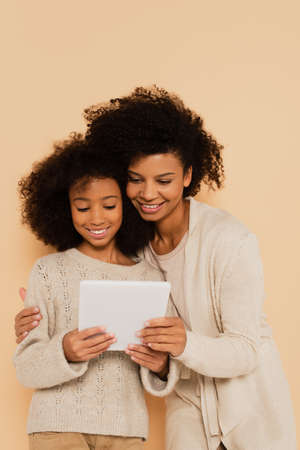 african american mother holding preteen daughter with tablet in hands isolated on beige
