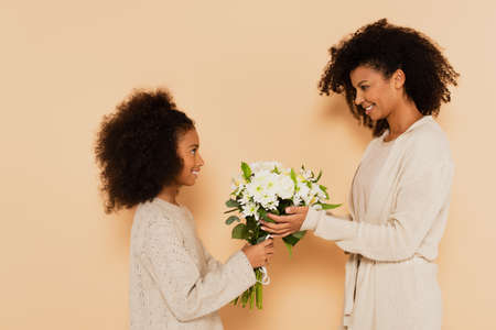 side view of african american preteen daughter and adult mother holding bouquet of daisies on beige backgroundの写真素材