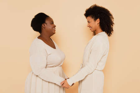 happy african american adult daughter and middle aged mother holding hands and looking at each other on beige backgroundの写真素材