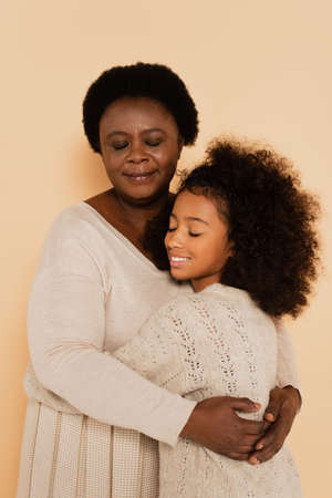 african american grandmother hugging granddaughter with closed eyes on beige background