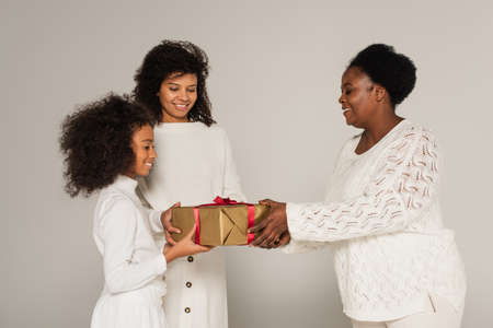 cheerful african american grandmother presenting gift box to daughter and granddaughter on gray...
