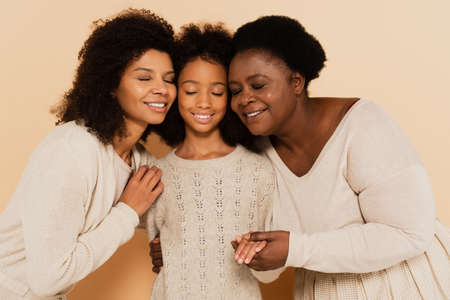african american daughter, granddaughter and grandmother hugging cheeks to cheeks and holding hands on beige backgroundの写真素材