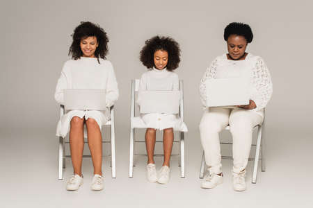 african american daughter, mother and granny sitting on chairs and looking at laptops on gray backgroundの写真素材