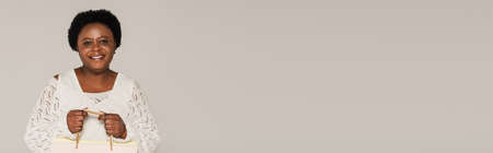 smiling african american middle aged woman in white clothes holding shopping bags isolated on gray, bannerの写真素材