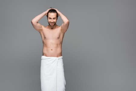 happy muscular man wrapped in towel adjusting hair isolated on gray