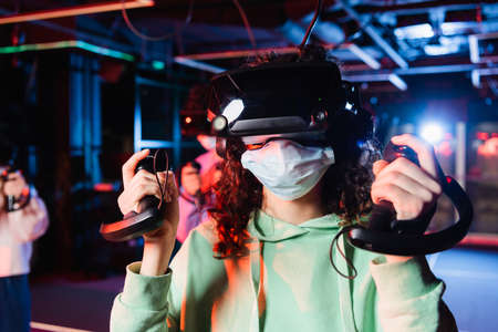 african american girl in medical mask and vr headset gaming near blurred friends