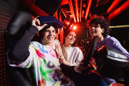 cheerful friends looking at camera near car racing simulator in game zone
