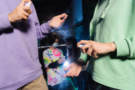 multiethnic friends applying hand sanitizer near blurred gamers in medical masks