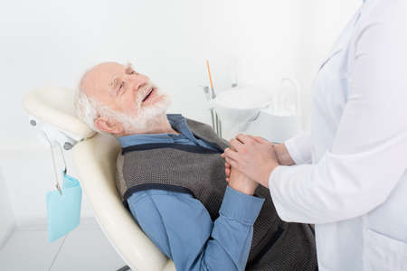 smiling senior patient lying in dental chair and holding hands of dentist in clinicの写真素材