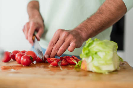 cropped view of man cutting cherry tomatoes near fresh lettuce in kitchenの写真素材