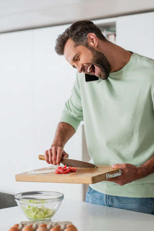 excited man laughing while preparing fresh salad and talking on smartphoneの写真素材
