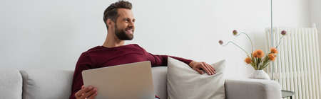 smiling man looking away while sitting on couch with laptop, bannerの写真素材