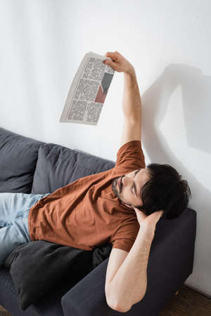 man waving with newspaper while lying on gray couch and suffering from heat