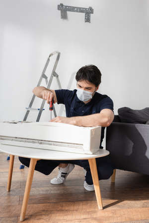 repairman in medical mask holding screwdriver while fixing air conditioner