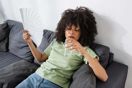 african american woman waving with hand fan while holding glass of water and sitting on gray couchの写真素材