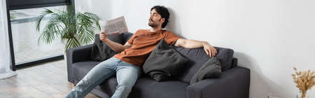 man waving with newspaper while lying on gray sofa and suffering from heat, bannerの写真素材