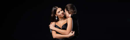 man in black blazer and white shirt kissing shoulder of elegant woman in silk dress and jewelry isolated on black, bannerの写真素材