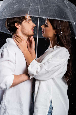 romantic young adult boyfriend and girlfriend standing in rain with umbrella on black background