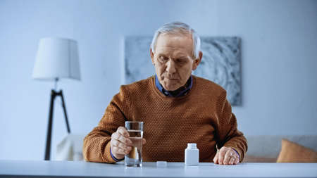 elderly man sitting with open medicine jar on table and holding glass of water in living roomの写真素材