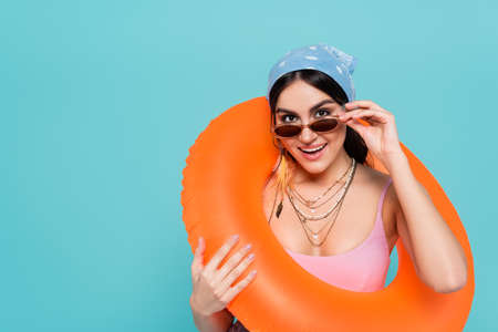 Cheerful woman in sunglasses holding swim ring isolated on blue