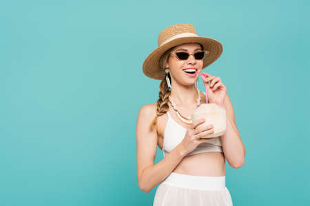 Cheerful woman in sunglasses holding cocktail in coconut isolated on blueの写真素材