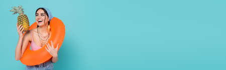 Cheerful woman with swim ring holding pineapple isolated on blue, banner