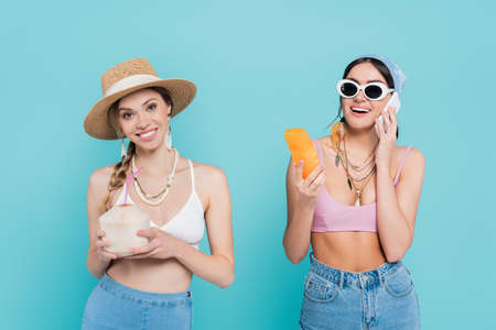 Smiling woman holding cocktail in coconut near friend with sunscreen talking on smartphone isolated on blueの写真素材