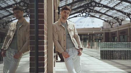 smiling man in beige jacket looking at camera and posing near building of shopping mall