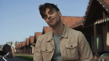 smiling man in beige jacket looking at camera near motorcycle