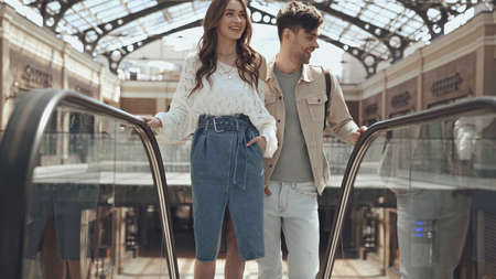 happy woman posing with hand in pocket near man on escalator in shopping mall
